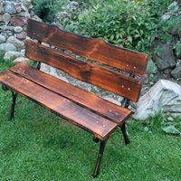 Rustykalna ławka ogrodowa z ciemnego drewna o nieregularnych krawędziach, osadzona na metalowych nogach, na tle zielonej trawy i kamiennego murku.