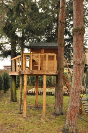 Drewniany domek na drzewie z balustradą, wsparty na palach, z drabinką, otoczony drzewami iglastymi na trawiastym terenie.