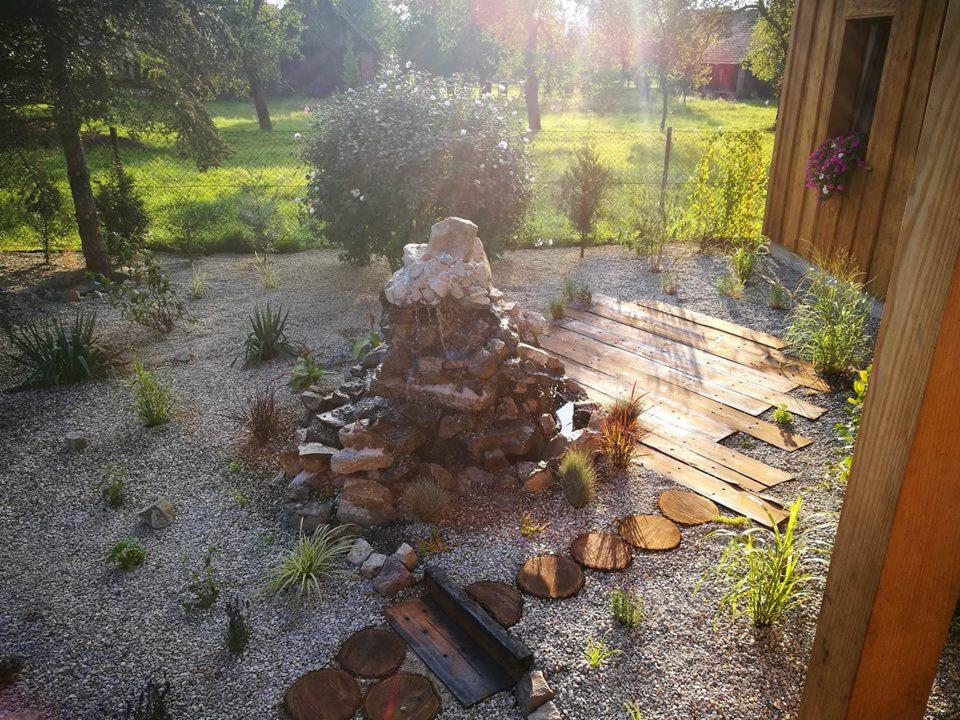 Ogród z kaskadowym wodospadem z kamieni, ścieżką z drewnianych bali i desek oraz rabatami żwirowymi w słoneczny dzień.