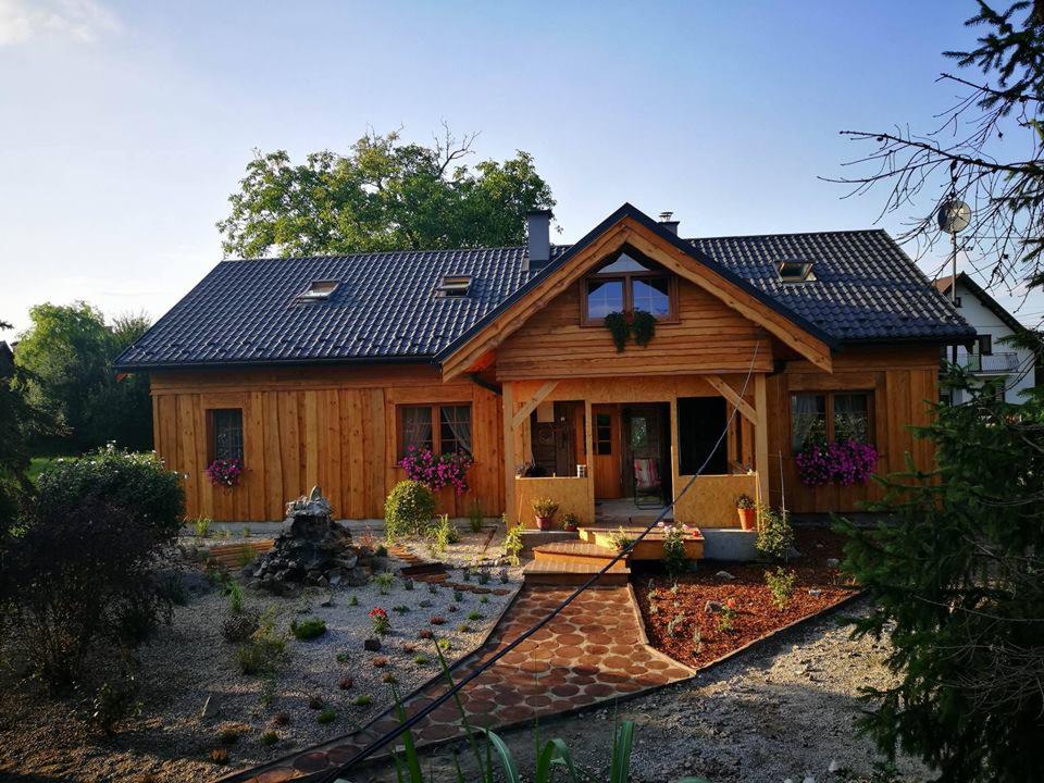 Drewniany dom jednorodzinny z ciemną dachówką, widok z ogrodu z kamienną fontanną i ścieżką z kostki. Kwiaty w donicach zdobią okna i wejście.