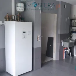 Biała jednostka wewnętrzna urządzenia grzewczego Panasonic z podłączonymi rurami, zamontowana w szarym pomieszczeniu z umywalką i kolektorami podłogowymi, widoczne logo firmy na ścianie.