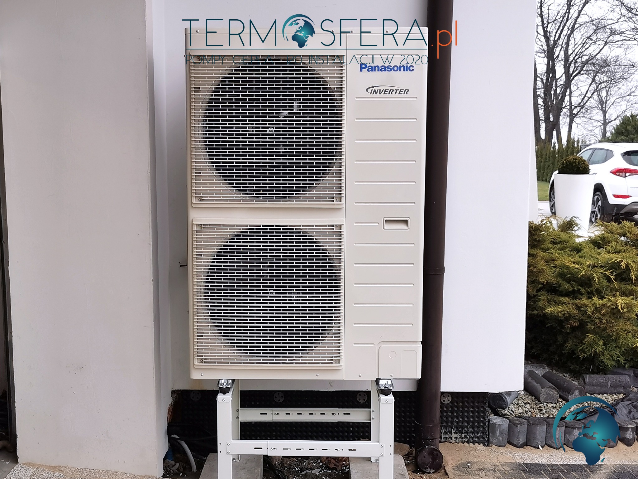 Zewnętrzna jednostka klimatyzacyjna Panasonic Inverter zamontowana na białej ścianie budynku z widocznym logo firmy Termosfera.pl, na tle ogrodu z białym samochodem.