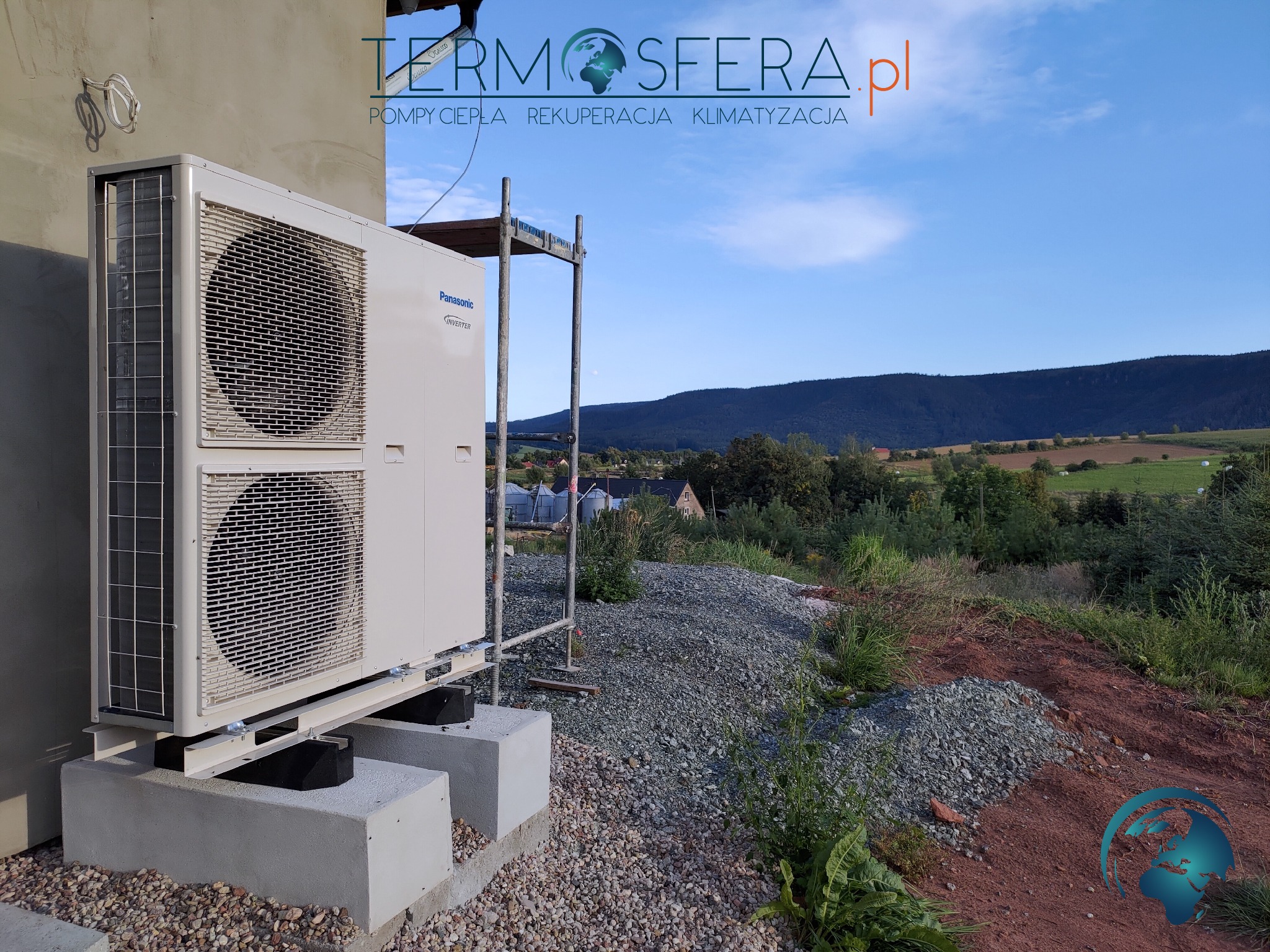 Zewnętrzna jednostka klimatyzacyjna Panasonic Inverter zamontowana na betonowych podstawach, z widocznym rusztowaniem w tle i krajobrazem górskim w oddali.