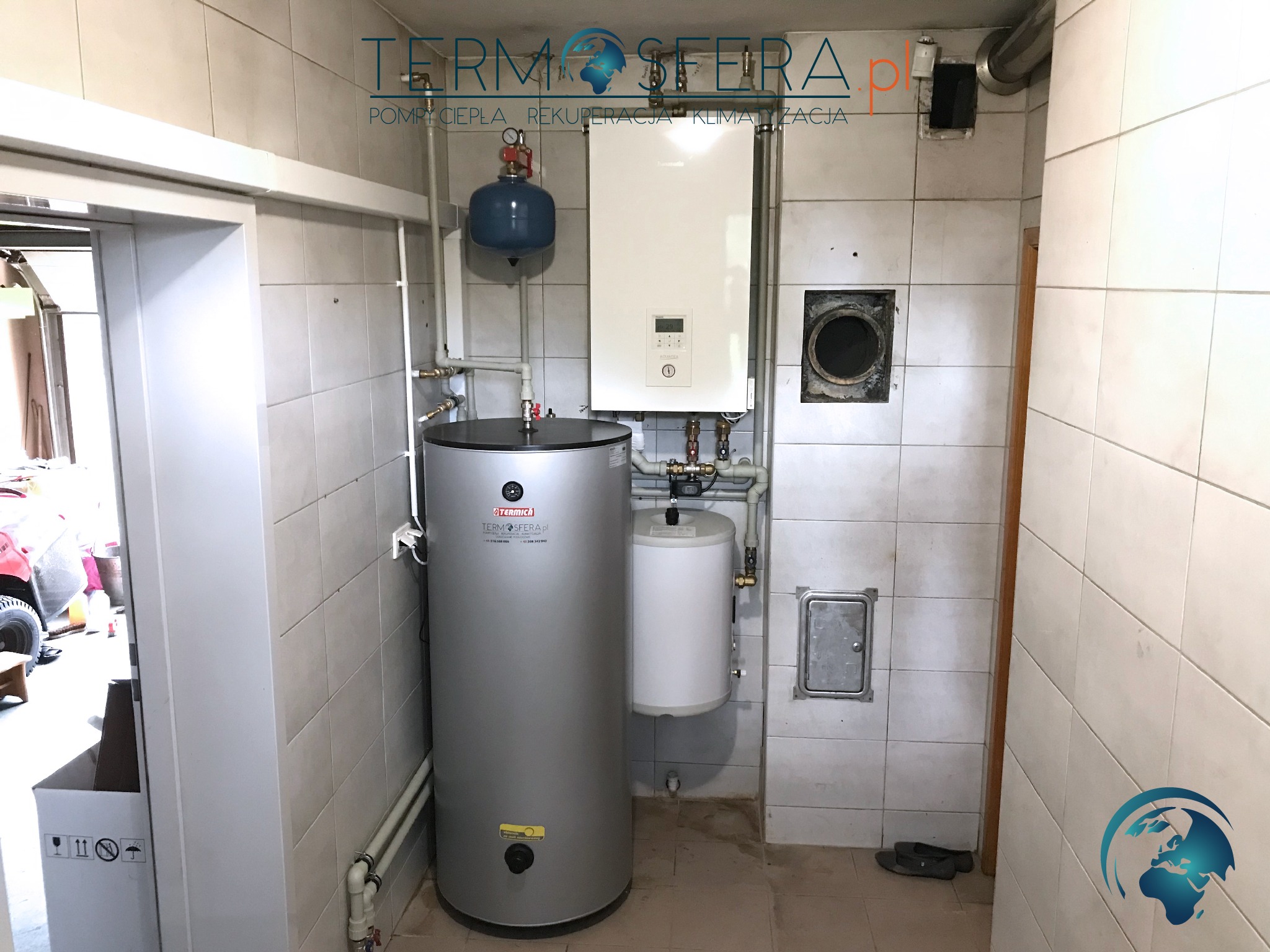 Instalacja grzewcza w kotłowni: zbiornik buforowy, wiszący bojler i piec grzewczy na tle białych, kwadratowych płytek ceramicznych. Widoczny manometr, rury miedziane i skrzynka rewizyjna.