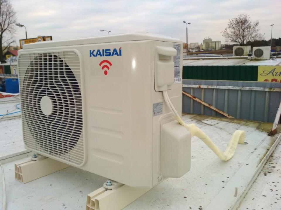 Zewnętrzna jednostka klimatyzacji Kaisai zamontowana na płaskim dachu, widoczne przewody w izolacji, w tle budynki miejskie i inne jednostki klimatyzacyjne.
