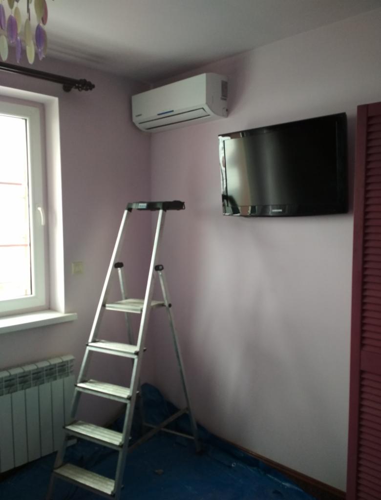 Biały klimatyzator ścienny zamontowany w pokoju z różowymi ścianami, obok wiszącego telewizora, z rozstawioną aluminiową drabiną na niebieskiej folii ochronnej.
