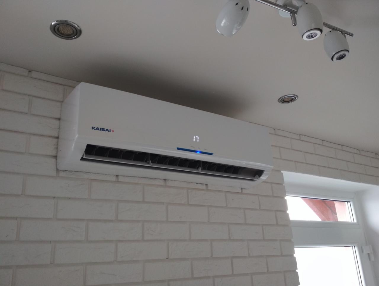 Biała jednostka klimatyzacyjna Kaisai zamontowana na ścianie z białej cegły w pomieszczeniu z oknem i nowoczesnym oświetleniem sufitowym.