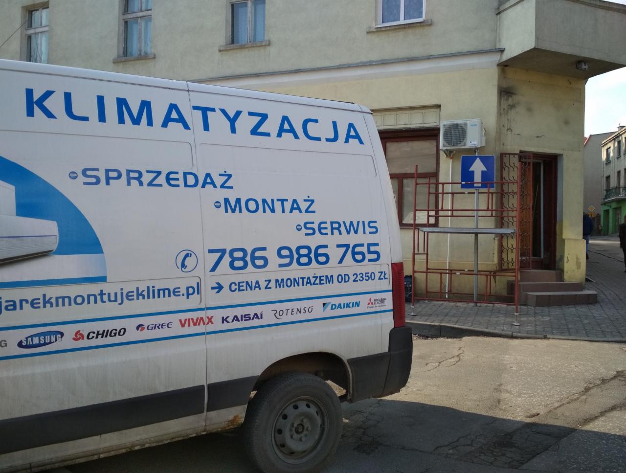 Biały samochód dostawczy z niebieskim napisem KLIMATYZACJA, numerem telefonu i logo producentów klimatyzatorów, zaparkowany na ulicy przed budynkiem z zamontowanym klimatyzatorem na ścianie.