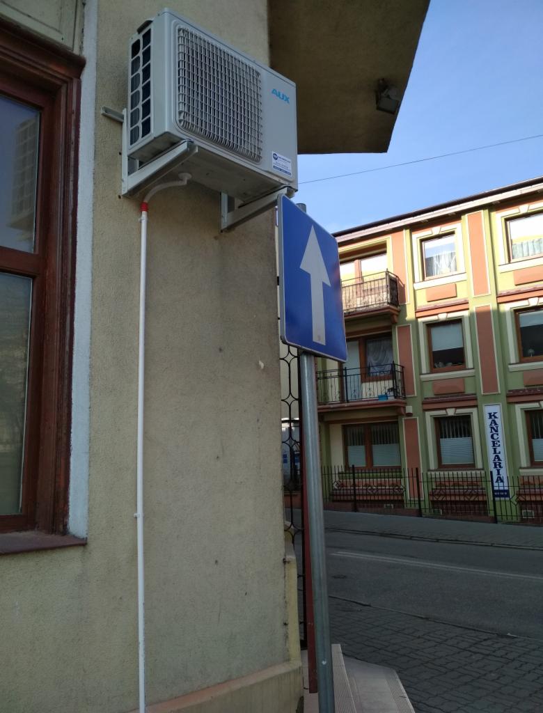 Zewnętrzna jednostka klimatyzacji AUX zamontowana na elewacji budynku, widoczny fragment rury odprowadzającej skropliny i znak drogowy.