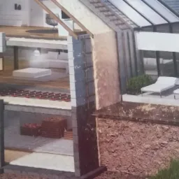 Przekrój domu w trakcie budowy ukazujący warstwy izolacyjne ścian oraz instalację ogrzewania podłogowego, z pracownikiem w kombinezonie natryskującym izolację na ścianę.