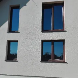 Elewacja budynku z beżowym tynkiem strukturalnym i czterema oknami z brązowymi ramami, na dole widoczny fragment ocieplenia fundamentu.