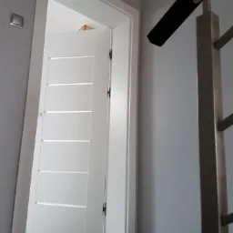 Białe drzwi wewnętrzne z poziomymi przeszkleniami, widoczne z perspektywy dolnej części schodów, z fragmentem metalowej balustrady po prawej stronie.