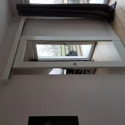 Okno dachowe uchylne z białą ramą, widok z dołu, szare zasłony w tle, drewniana podłoga.