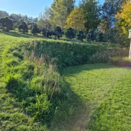 Ukształtowany teren zielony z trawnikiem, skarpa z wysoką trawą ozdobną, ogrodzenie z tujami w tle i kamienna altana z ułożonym drewnem.