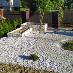 Nowo urządzony ogród z ścieżką z kostki granitowej ułożoną w okrąg wokół trawnika, rabatami z kamienia, drzewem i tujami wzdłuż drewnianego ogrodzenia.