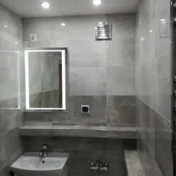 Wykończona łazienka w odcieniach szarości z podświetlanym lustrem, wentylatorem na ścianie i armaturą. Widoczne płytki o zróżnicowanej fakturze i oświetlenie punktowe.