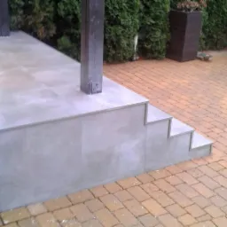 Wykończony taras z szarymi płytkami i schodami, widok z zewnątrz, na pierwszym planie kostka brukowa w odcieniach brązu, w tle zieleń i fragment samochodu.