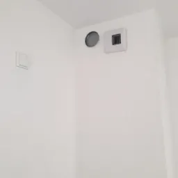 Biała ściana w stanie deweloperskim z widocznymi otworami wentylacyjnymi i włącznikiem światła, zagruntowana powierzchnia gotowa do dalszych prac wykończeniowych.