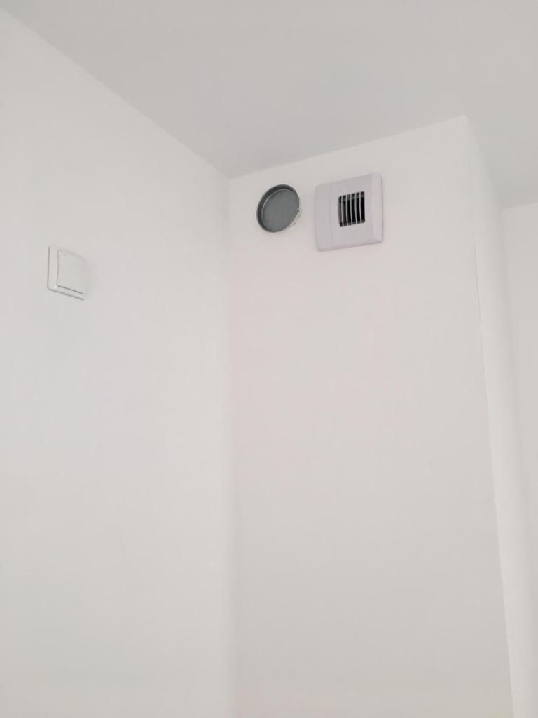 Biała ściana w stanie deweloperskim z widocznymi otworami wentylacyjnymi i włącznikiem światła, zagruntowana powierzchnia gotowa do dalszych prac wykończeniowych.