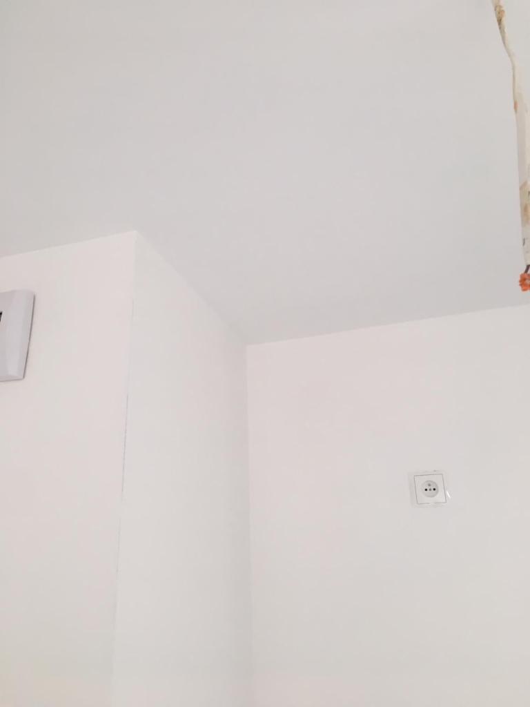 Wnętrze w stanie deweloperskim: białe, zagruntowane ściany i sufit, widoczne gniazdko elektryczne i fragment instalacji zabezpieczonej folią.