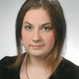 Portret kobiety w czarnej bluzce i ze złotym łańcuszkiem, uśmiechnięta, na jasnym tle.