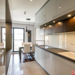 Deco Nova - Nowoczesna kuchnia z białymi szafkami, ciemnym blatem i oświetleniem podszafkowym, widok na stół z krzesłami i lampami wiszącymi.