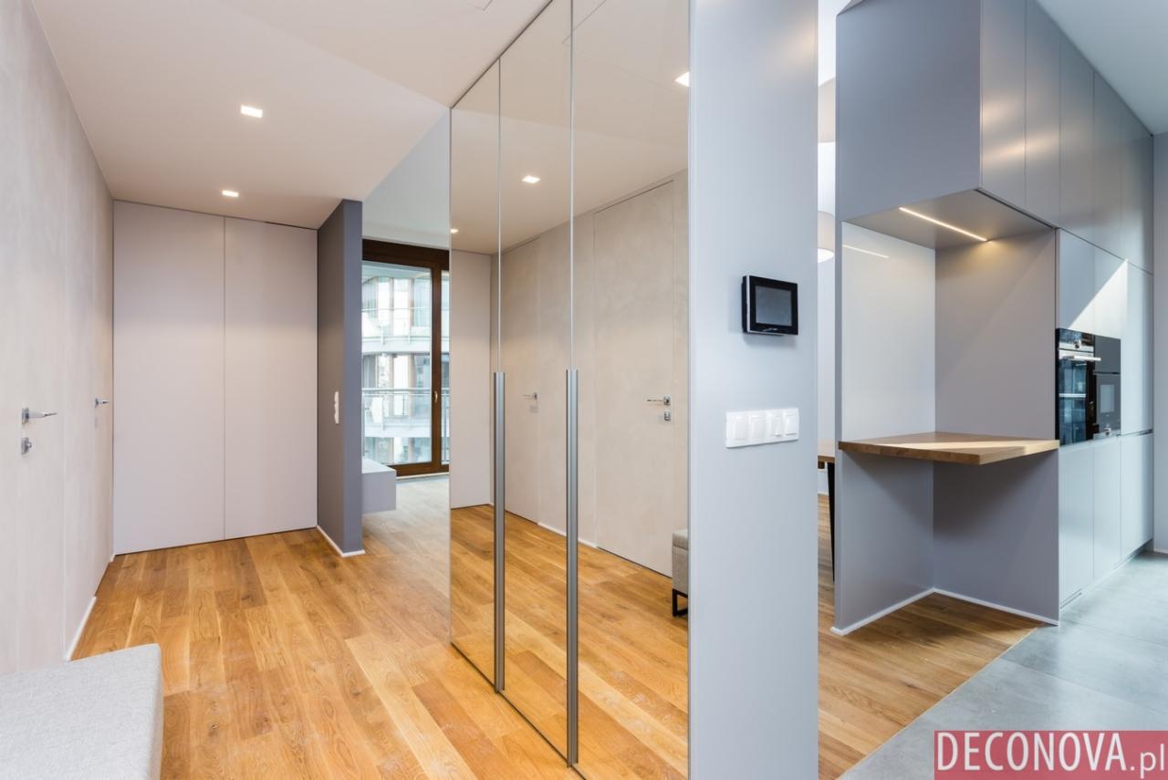 Nowoczesne wnętrze mieszkania z drewnianą podłogą, lustrzanymi drzwiami szafy wnękowej, szarymi ścianami i zabudową kuchenną z drewnianym blatem