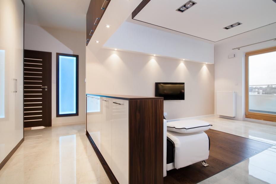 Nowoczesne wnętrze z błyszczącą, marmurową podłogą, niską zabudową meblową z frontami w bieli i ciemnym drewnie, telewizorem na ścianie i minimalistycznym fotelem z poduszką.