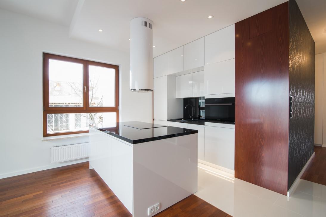 Nowoczesna kuchnia z białą wyspą i czarnym blatem, cylindryczny okap, drewniana podłoga i fornir na ścianie