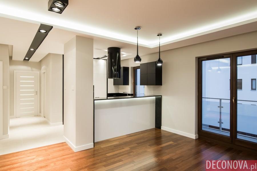 Nowoczesne wnętrze mieszkania z drewnianą podłogą, białymi ścianami i aneksem kuchennym z czarnymi szafkami, oświetleniem punktowym i barkiem z podświetleniem LED.