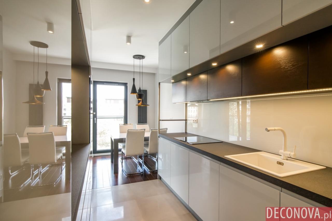 Nowoczesna kuchnia z białymi szafkami, ciemnym blatem i oświetleniem podszafkowym, widok na stół z krzesłami i lampami wiszącymi.