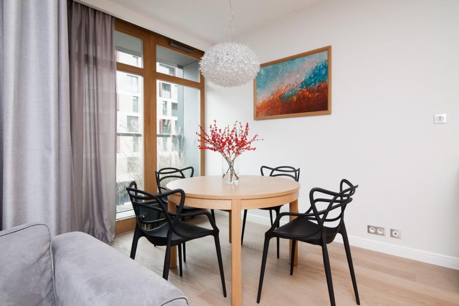 Jasny salon z okrągłym drewnianym stołem otoczonym czarnymi krzesłami o nowoczesnym designie, czerwone gałązki w wazonie na stole, abstrakcyjny obraz na ścianie i designerska lampa wisząca.