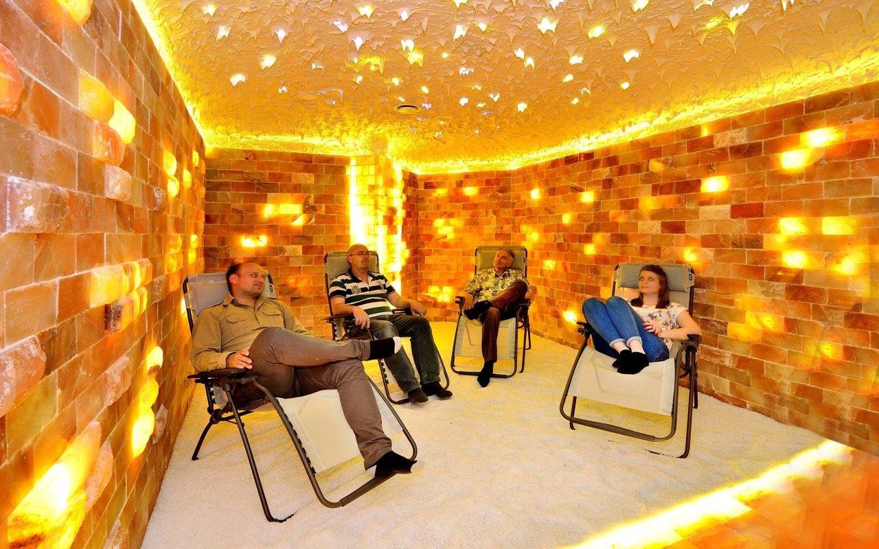 Wnętrze sauny solnej z czterema osobami relaksującymi się w leżakach na białym podłożu, ściany i sufit wyłożone cegłami solnymi podświetlonymi od wewnątrz.