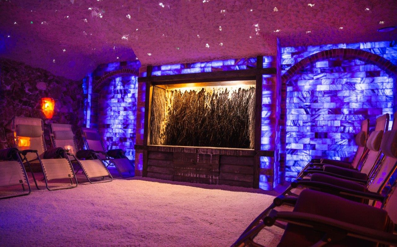 Wnętrze groty solnej z leżakami, podświetlonymi ścianami z bloków soli i tężnią solankową w drewnianej obudowie.