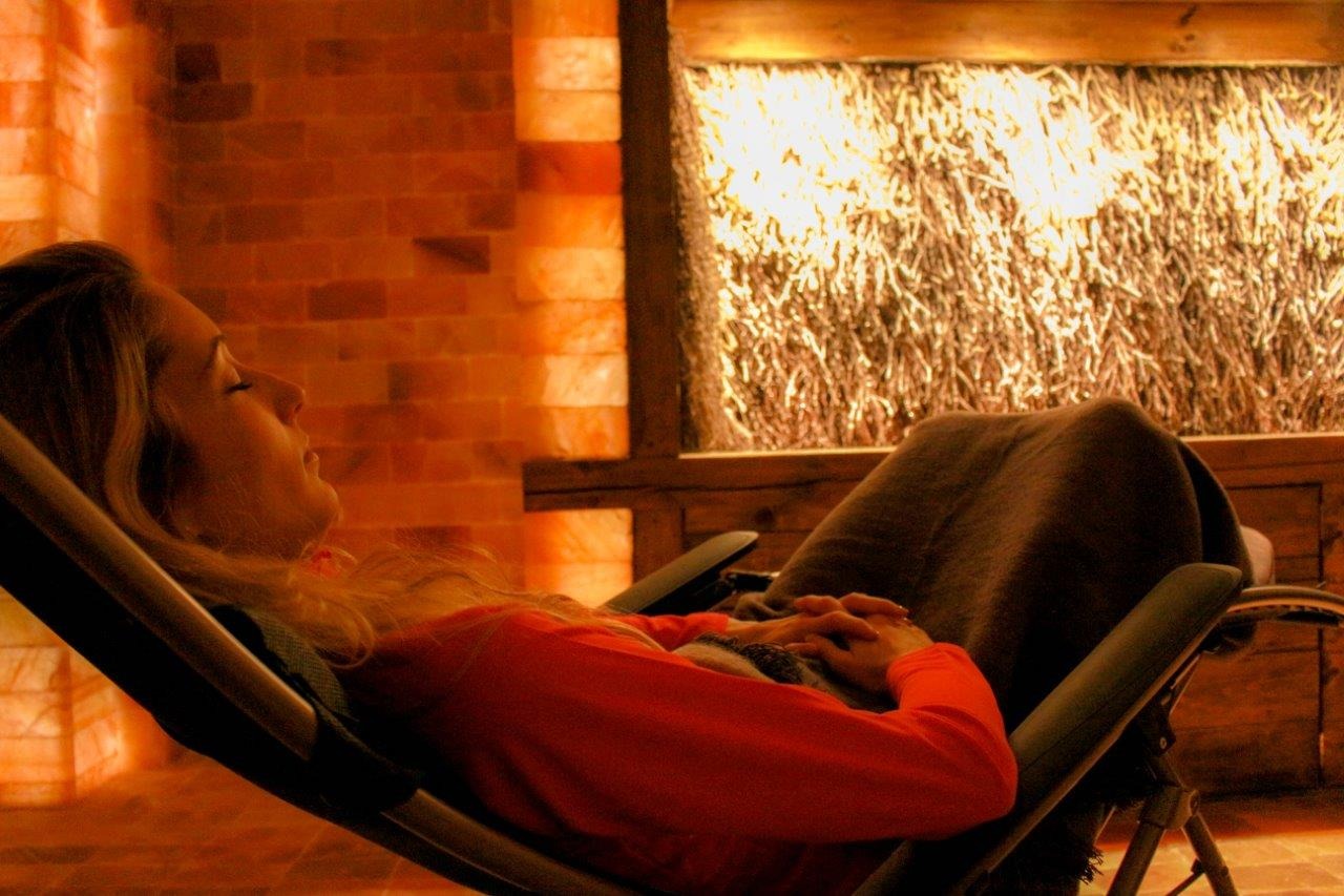 Kobieta relaksuje się w fotelu w pomieszczeniu wyłożonym cegłami solnymi, z widoczną ścianą z tężni solankowej w tle.