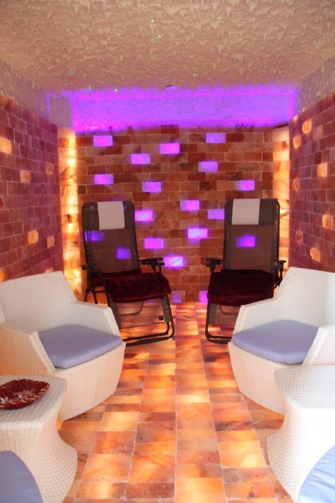 Wnętrze sauny solnej z dwoma wiklinowymi fotelami i dwoma leżakami, ściany i podłoga z bloków soli, oświetlenie LED w kolorze fioletowym.