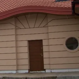 Budynek z elewacją w beżowych pasach, brązowe drzwi wejściowe i okrągłe okno, dach z dachówką. Widoczna linia styku elewacji z chodnikiem.