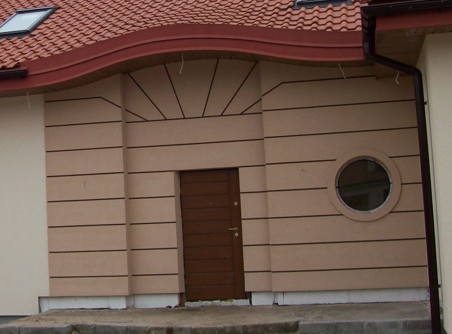 Budynek z elewacją w beżowych pasach, brązowe drzwi wejściowe i okrągłe okno, dach z dachówką. Widoczna linia styku elewacji z chodnikiem.