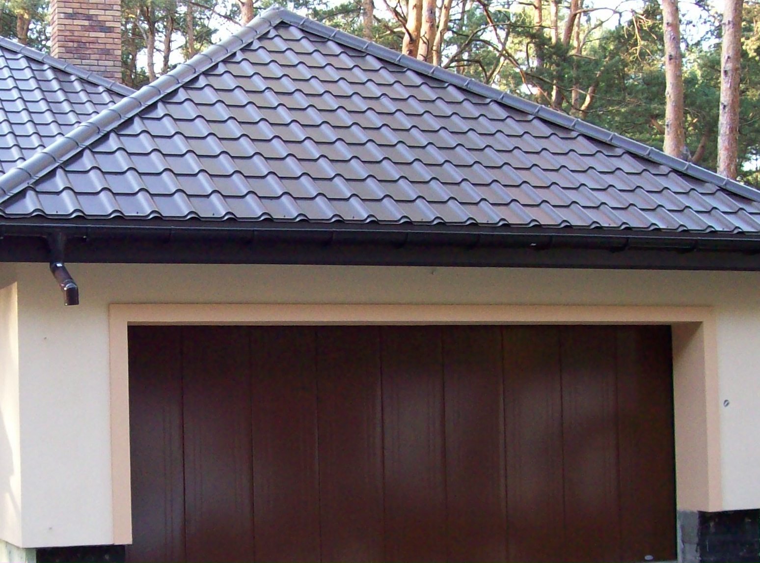 Dom z brązową bramą garażową segmentową i ciemną dachówką, widok z zewnątrz w słoneczny dzień.