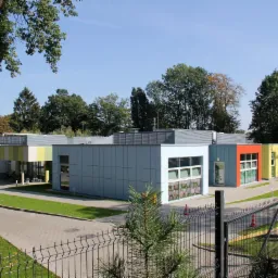 Nowoczesny, parterowy budynek o modułowej konstrukcji z płaskim dachem i kolorowymi elementami fasady, otoczony zielenią i ogrodzeniem.