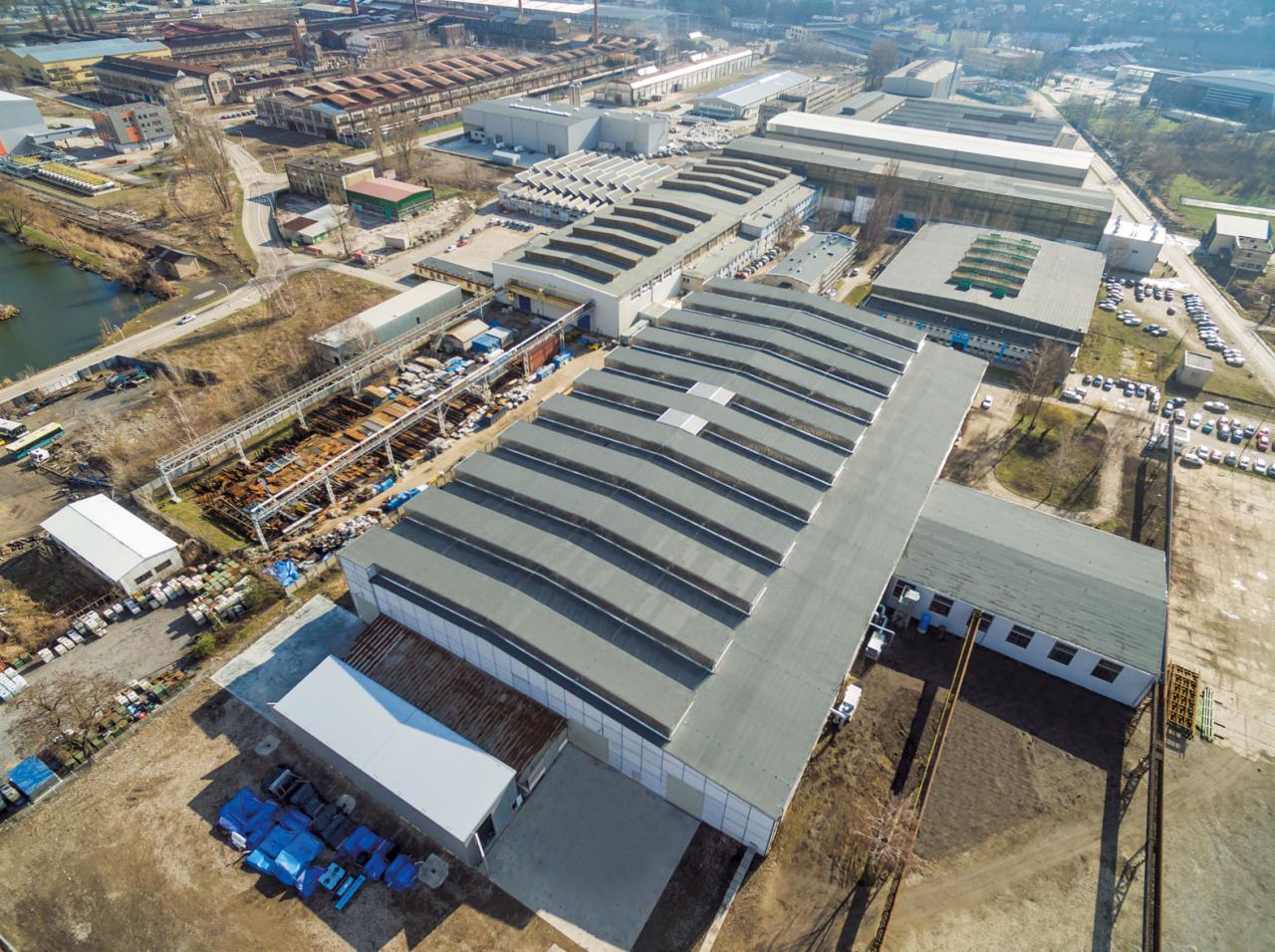 Widok z lotu ptaka na rozległy kompleks przemysłowy z halami o charakterystycznych, falistych dachach, placem składowym z elementami konstrukcyjnymi i otaczającą infrastrukturą.