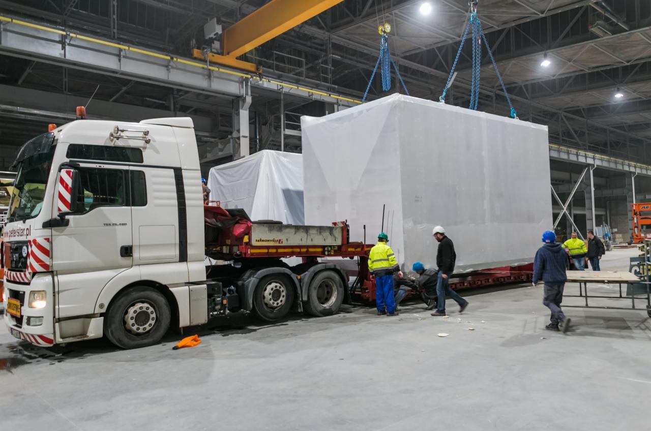 Transport modułu w folii ochronnej na naczepie niskopodwoziowej w hali produkcyjnej, z wykorzystaniem suwnicy, obsługa w kaskach i odblaskowych kamizelkach zabezpiecza ładunek.