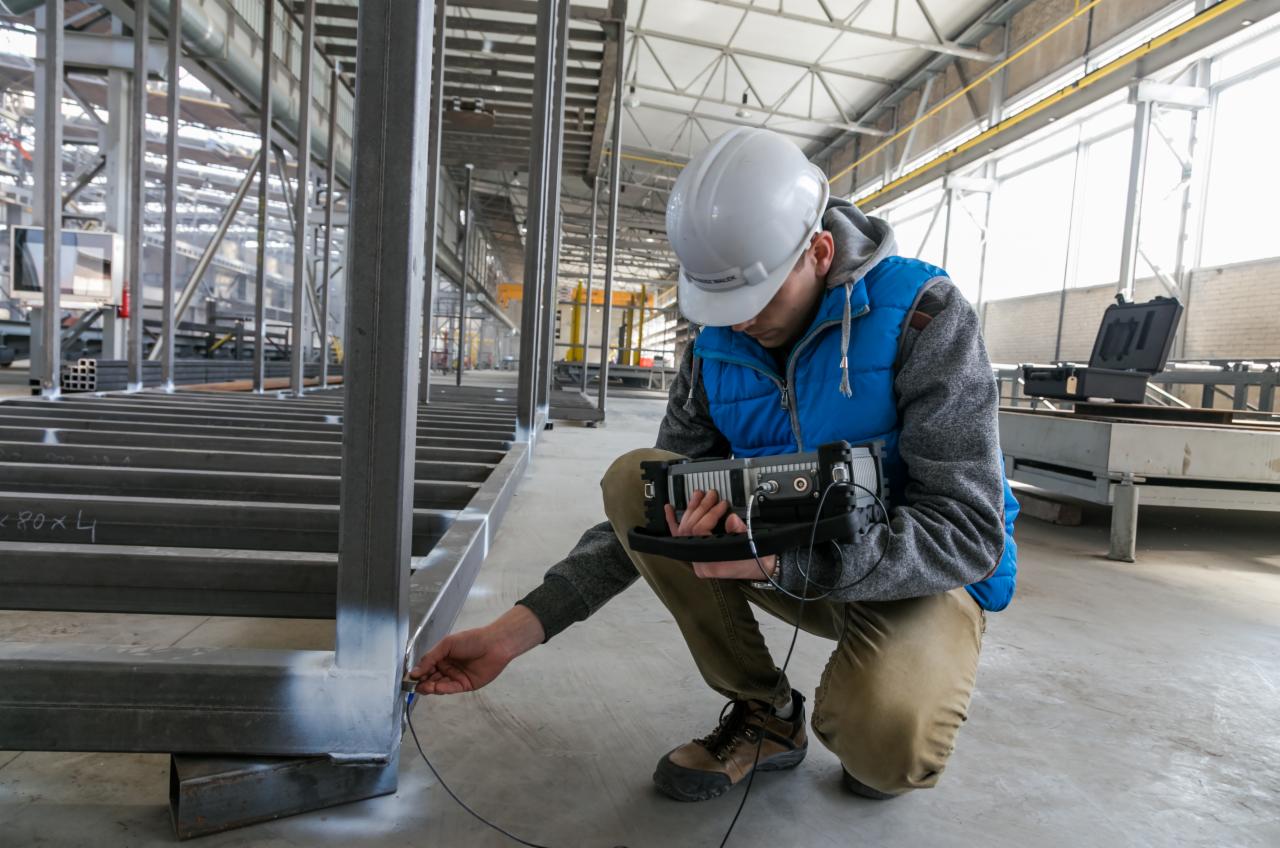 Pracownik w kasku kontroluje spawy konstrukcji stalowej za pomocą specjalistycznego urządzenia pomiarowego w hali produkcyjnej.