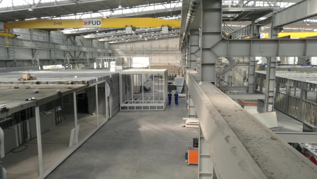 Wnętrze hali produkcyjnej z widocznymi elementami konstrukcyjnymi, w tym szkieletami przyszłych domów, suwnicą FUD pod sufitem i pracownikami w niebieskich kombinezonach.