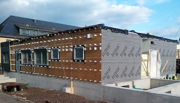 Budynek w trakcie budowy, widoczne ściany z izolacją termiczną i elementami konstrukcyjnymi, fundament betonowy, częściowo otynkowany, w tle budynek z ciemnym dachem.