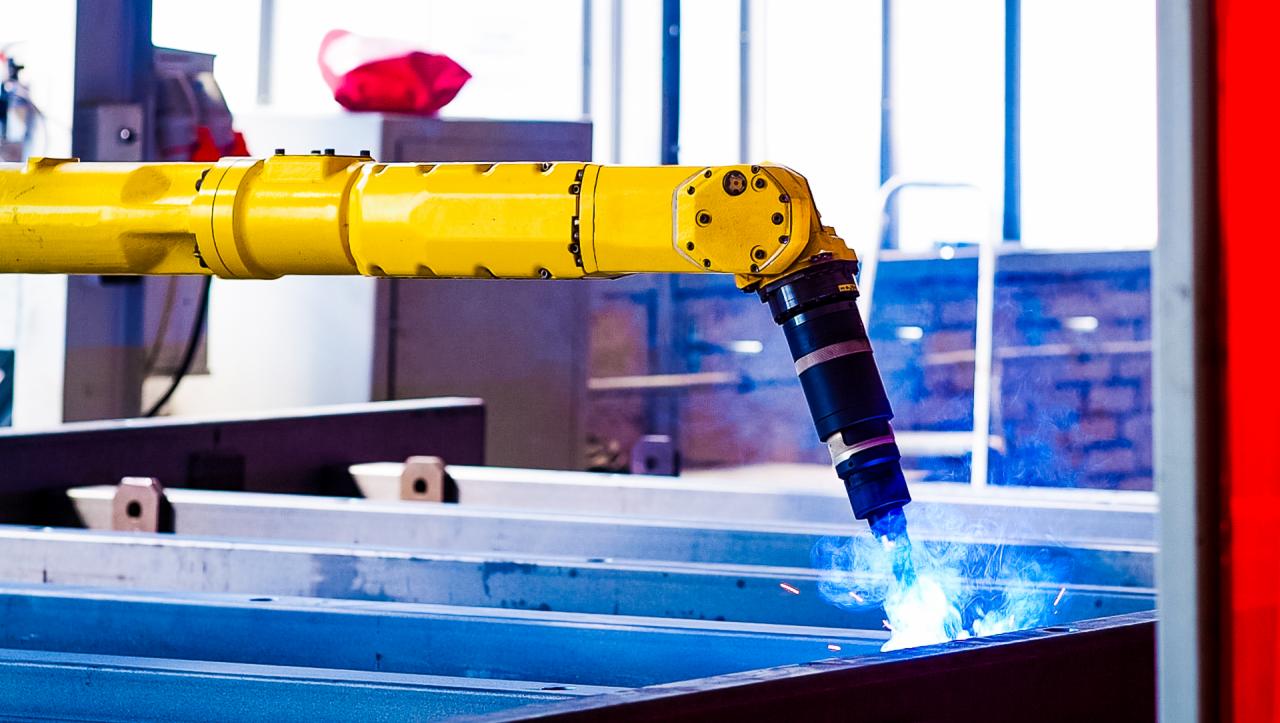 Żółty robot spawalniczy w trakcie pracy, spawający metalowe profile w hali produkcyjnej, widoczne iskry i niebieskie światło spawania.