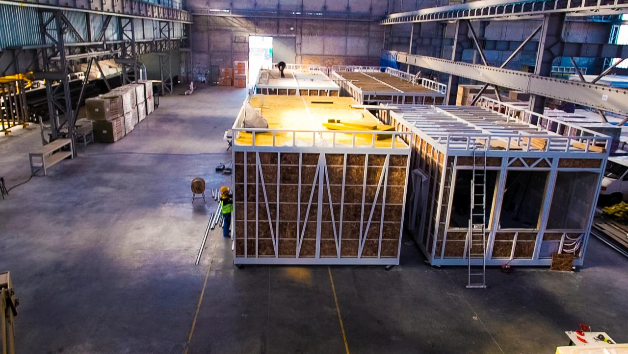 Wnętrze hali produkcyjnej z kilkoma modułami domów w trakcie budowy, widoczni pracownicy w odblaskowych kamizelkach i kaskach, konstrukcja szkieletowa z izolacją, ujęcie z góry.