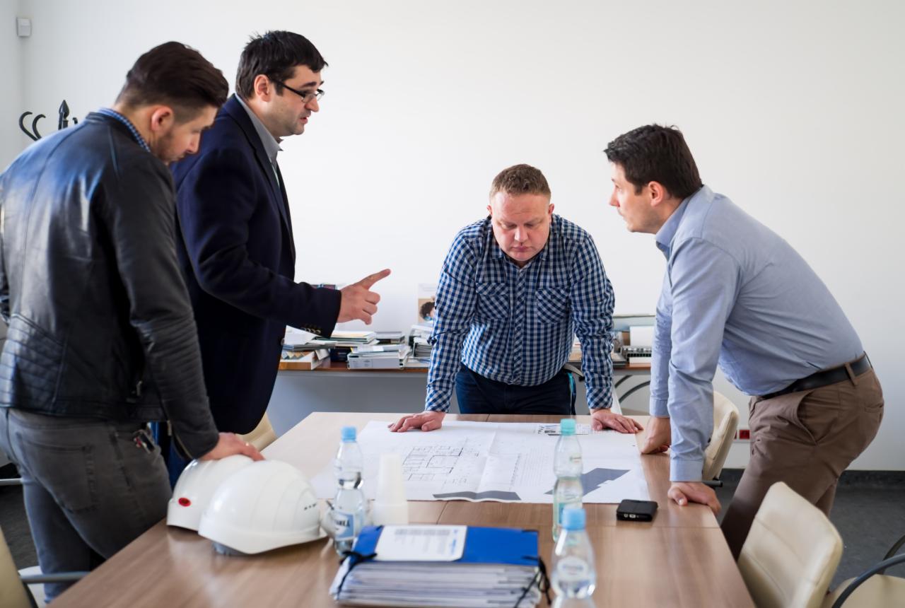 Czterech mężczyzn w strojach roboczych analizuje plan architektoniczny rozłożony na stole w jasnym biurze, widoczne kaski ochronne i butelki wody.