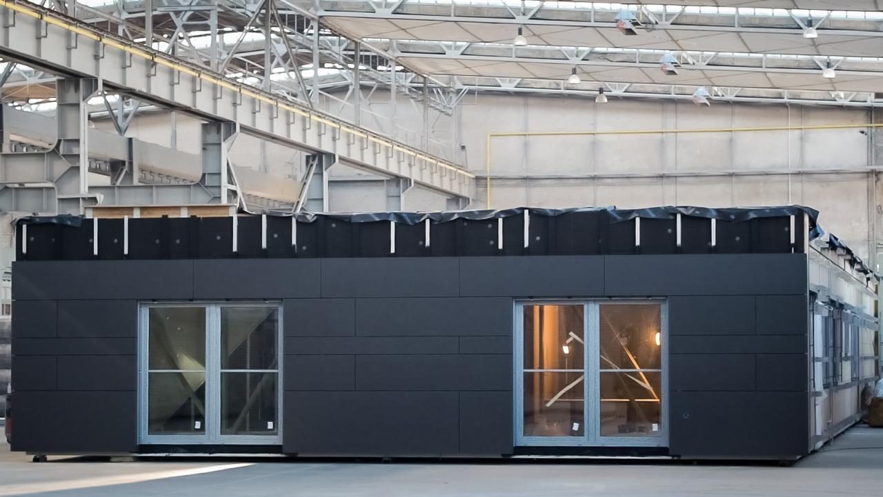 Ciemnoszary moduł domu z oknami, w hali produkcyjnej, z widocznym fragmentem konstrukcji dachu i izolacji zewnętrznej.