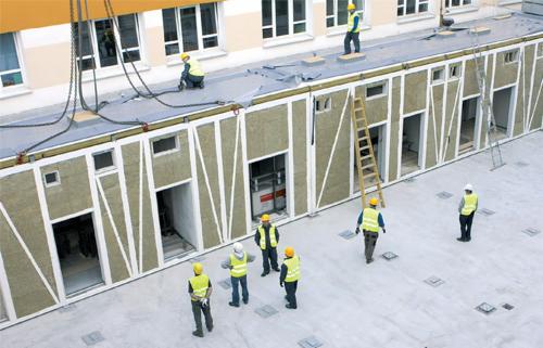 Montaż segmentów budynku z izolacją, widoczni pracownicy w żółtych kamizelkach i kaskach, drabina oparta o ścianę, żuraw podtrzymujący element konstrukcyjny.